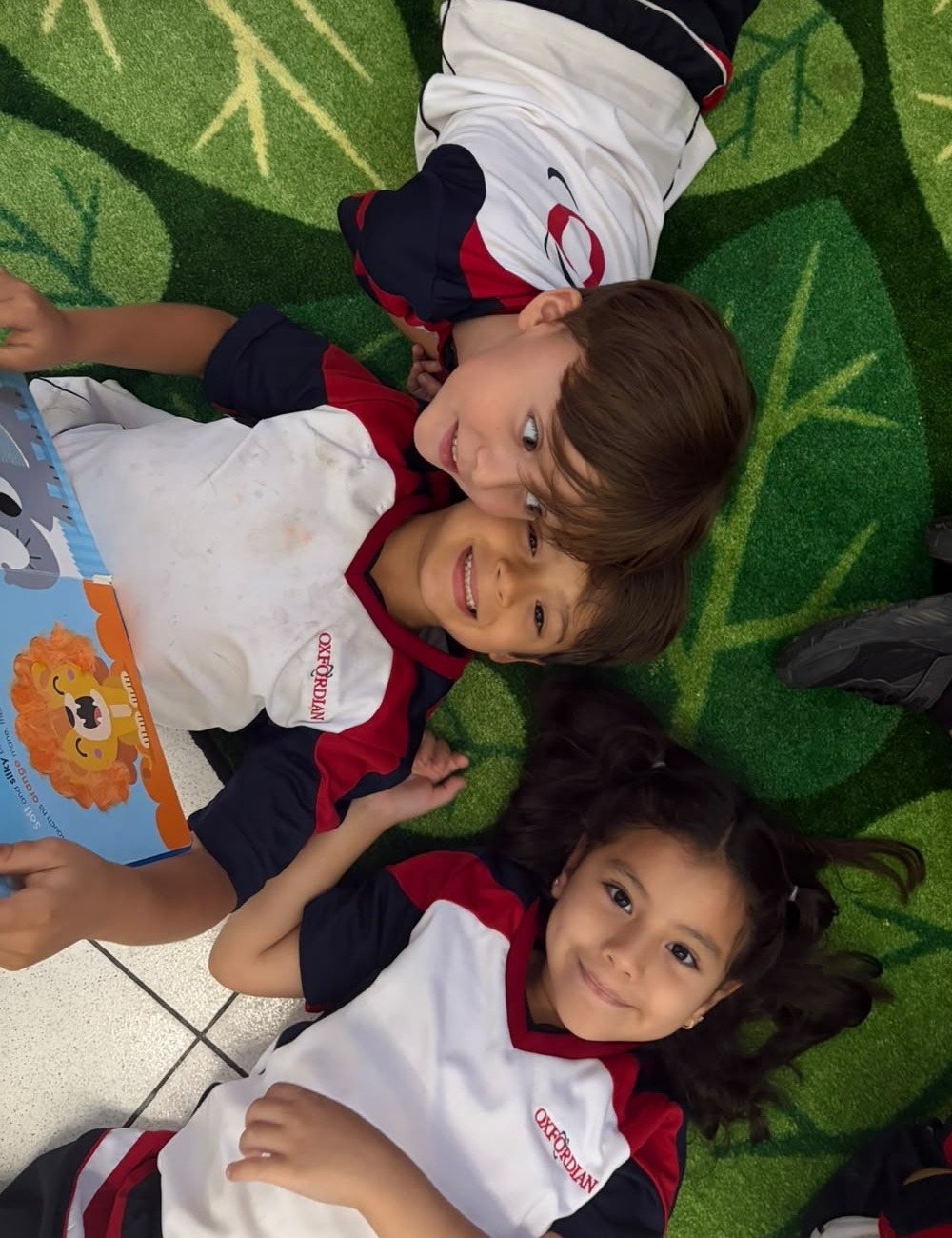 Three Kínder students lying on their classroom reading mat, one holding an open picture book