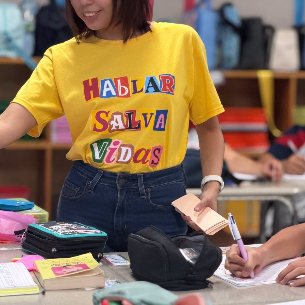 A member of staff in a t-shirt reading Hablar Salva Vidas hands out cards during a classroom session.