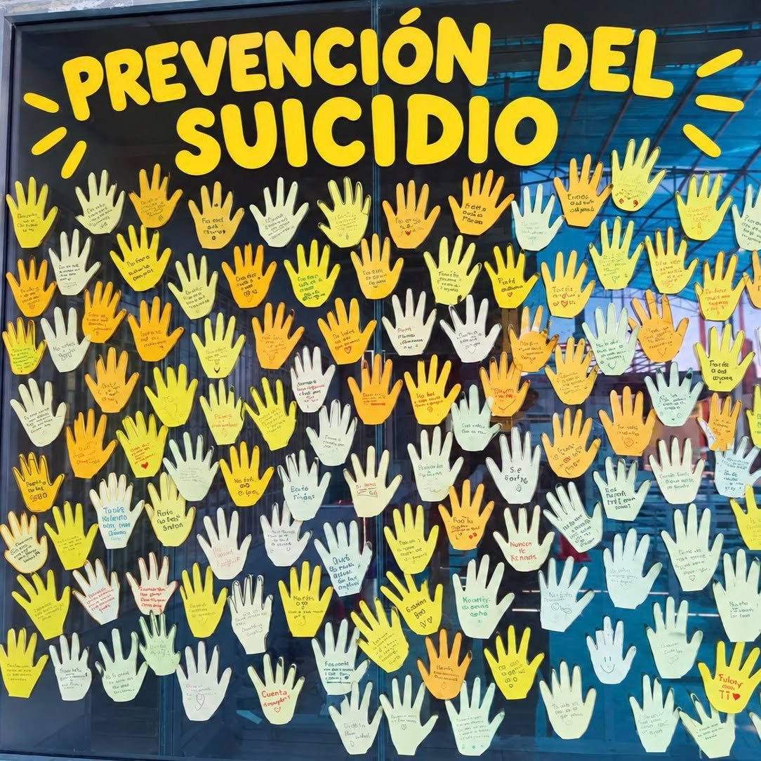 A school window under the heading Prevencion del Suicidio, covered with paper hands carrying handwritten messages of encouragement.