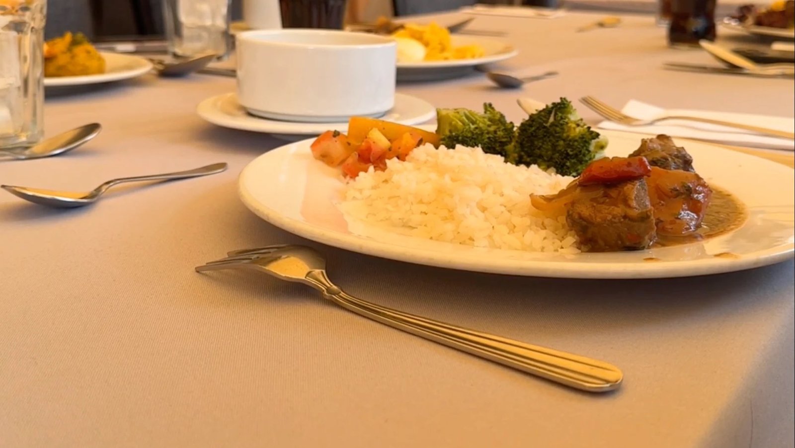 A cafetería lunch laid out on a set table: white rice, beef in tomato sauce, broccoli and diced vegetables on a plate, with cutlery and a water glass beside it.